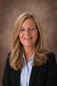 Wendy | Aspen Associates Group Head-and-shoulders portrait of a smiling woman with blond hair, wearing a light blue blouse and black blazer against a brown studio backdrop.